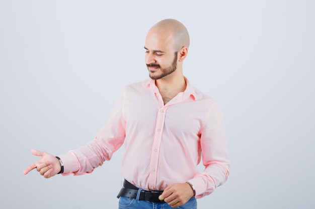 Foto gratuita giovane maschio che indica da parte in camicia, jeans e sembra gioioso. vista frontale.