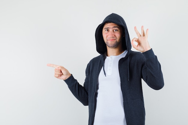 Young male pointing aside, showing ok gesture in t-shirt, jacket and looking confident , front view.