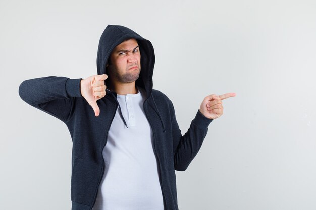 Young male pointing aside, showing thumb down in t-shirt, jacket and looking displeased , front view.