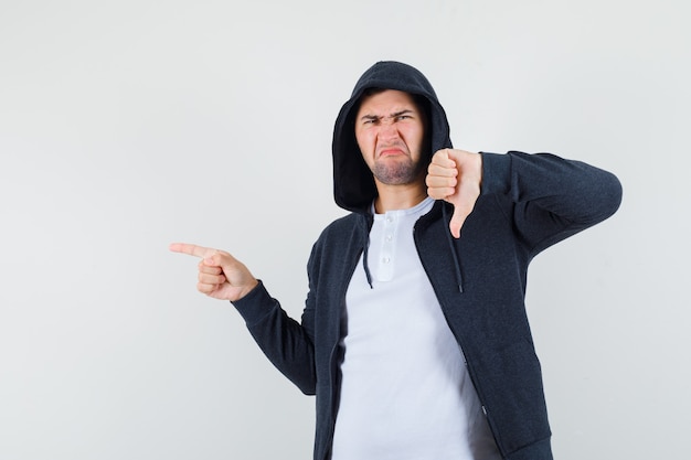 Young male pointing aside, showing thumb down in t-shirt, jacket and looking displeased , front view.