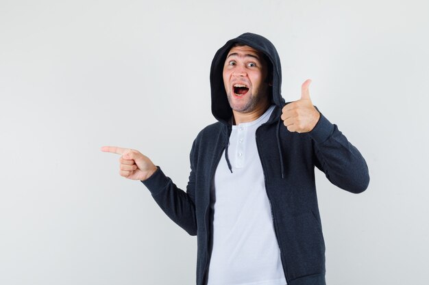 Young male pointing aside, showing thumb up in t-shirt, jacket and looking glad. front view.