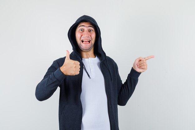 Young male pointing aside, showing thumb up in t-shirt, jacket and looking glad , front view.