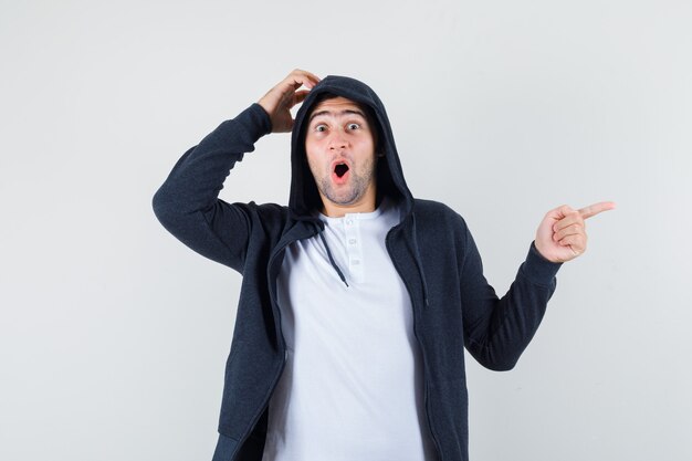 Young male pointing aside while scratching head in t-shirt, jacket and looking amazed. front view.