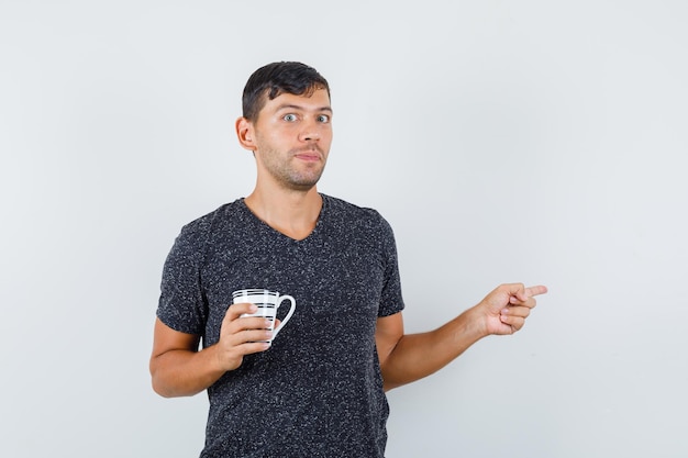 Young male pointing away while holding his cup in black t-shirt and looking focused , front view.