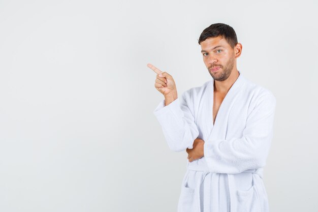 Young male pointing away in white bathrobe and looking confident. front view.
