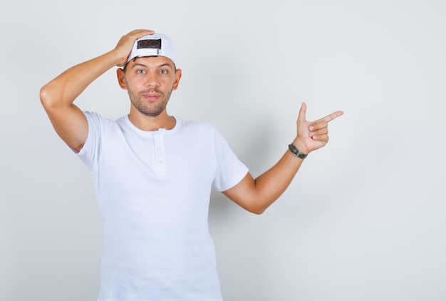 Young male pointing away with hand on head in white t-shirt, cap, front view.
