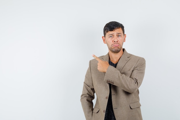 Young male pointing back in grayish brown jacket and looking disappointed , front view. space for text