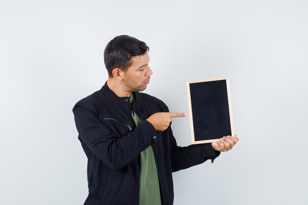 Young male pointing at blackboard in t-shirt, jacket and looking focused , front view.