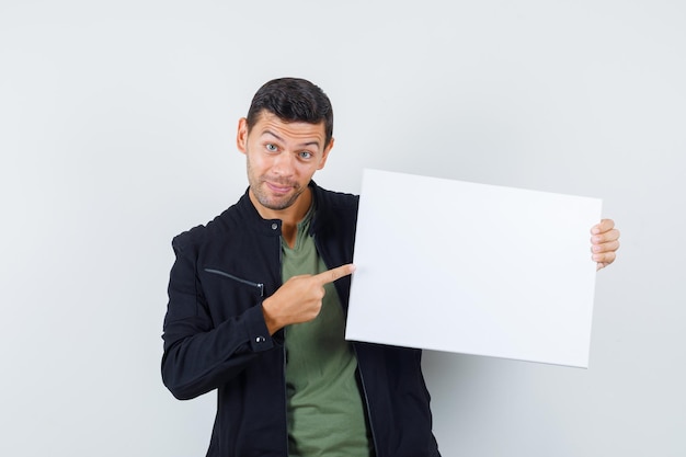 Young male pointing at blank canvas in t-shirt, jacket and looking cheerful , front view.