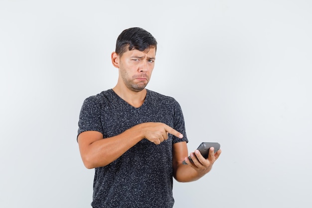 Young male pointing to calculator in black t-shirt and looking upset , front view.