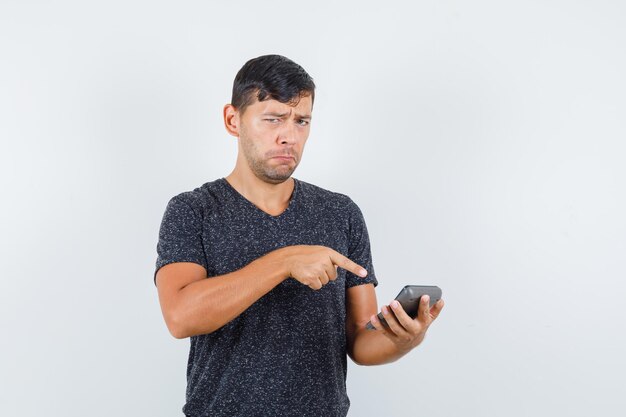Молодой мужчина, указывая на калькулятор в черной футболке и глядя расстроенным, вид спереди.