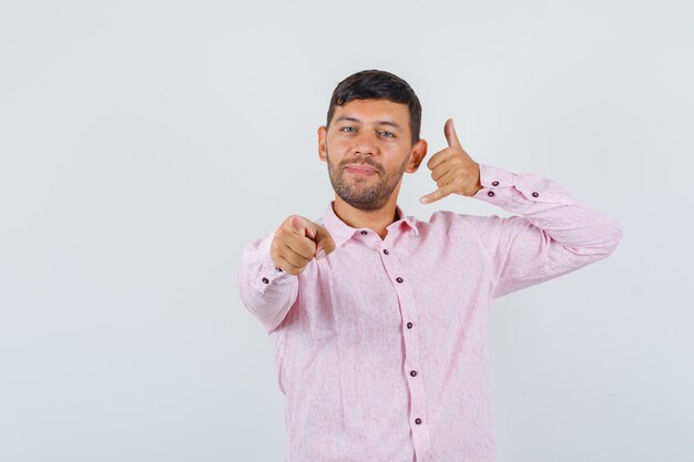 Young male pointing at camera with phone gesture in pink shirt and looking cheery. front view.