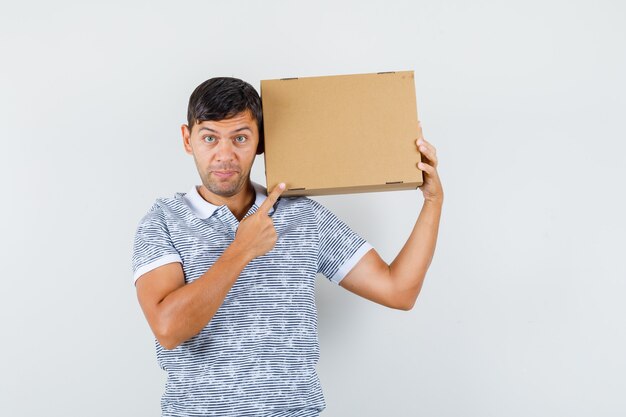 Young male pointing at cardboard box in t-shirt and looking cheerful