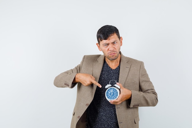 Young male pointing clock in grayish brown jacket and looking displeased , front view.