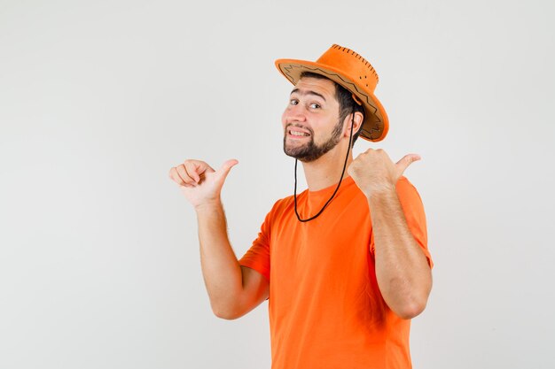 Young male pointing double thumbs up to the side in orange t-shirt, hat and looking merry. front view.