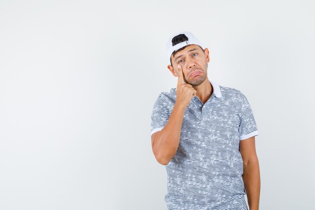 Young male pointing at eye in t-shirt and cap and looking fatigued 