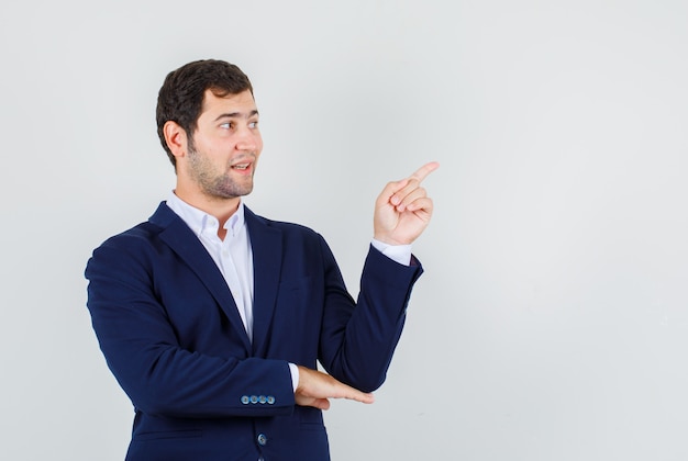 Young male pointing finger away in suit and looking glad. front view.