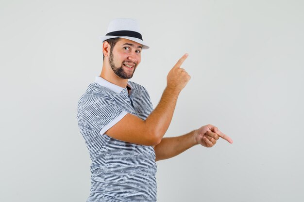 Young male pointing fingers up and down in t-shirt, hat and looking cheerful. front view.