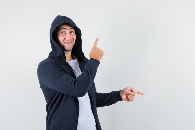 Young male pointing fingers up and down in t-shirt, jacket and looking cheerful , front view.