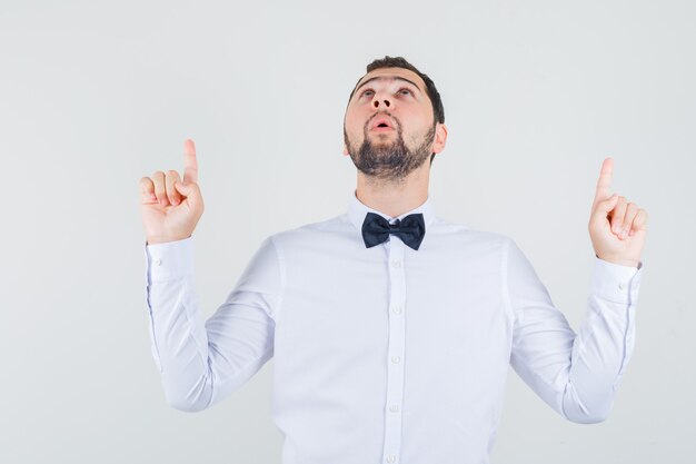 Young male pointing fingers up in white shirt and looking grateful. front view.