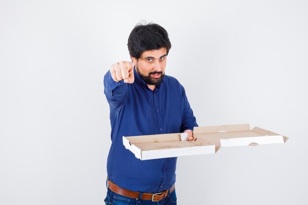 Young male pointing at front while holding pizza box in shirt, jeans and looking confident , front view.