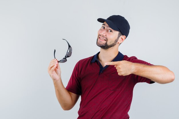 Young male pointing at glasses in t-shirt,black glasses and looking cheerful , front view.