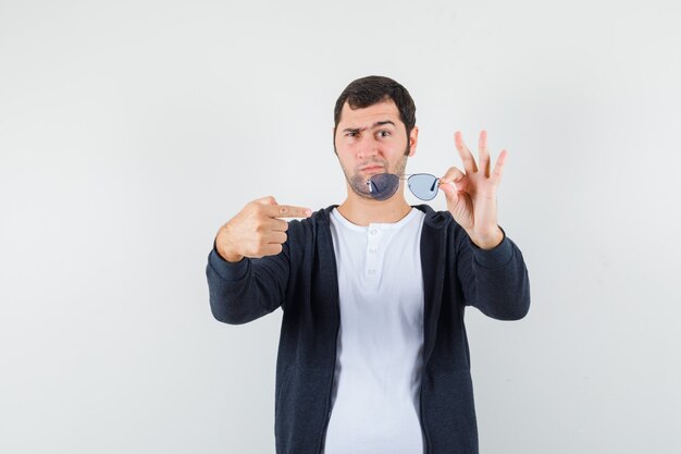 Young male pointing at glasses in t-shirt, jacket and looking pensive , front view.