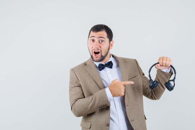 Young male pointing at headphones in suit and looking positive , front view.