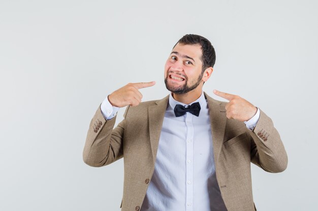 Young male pointing at his smile in suit and looking beloved , front view.