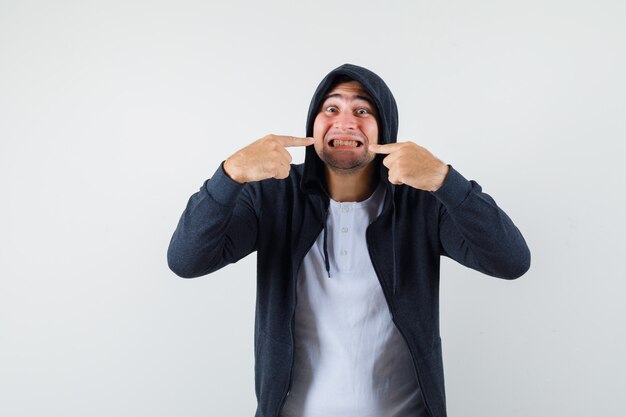 Young male pointing at his teeth in t-shirt, jacket and looking cheery. front view.