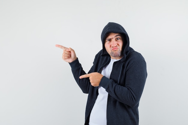 Young male pointing to the left side in t-shirt, jacket and looking pensive. front view.