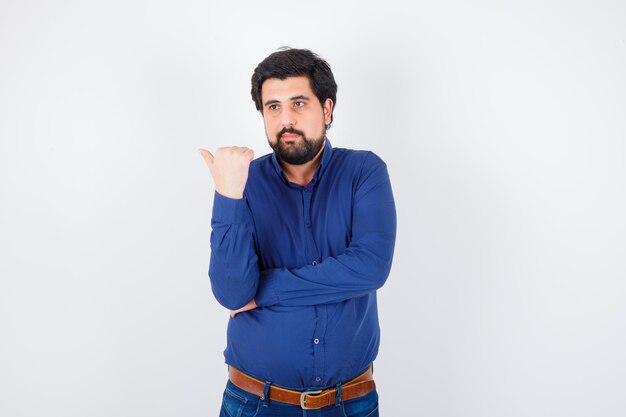 Young male pointing to the left side with thumb in shirt, jeans and looking confident , front view.