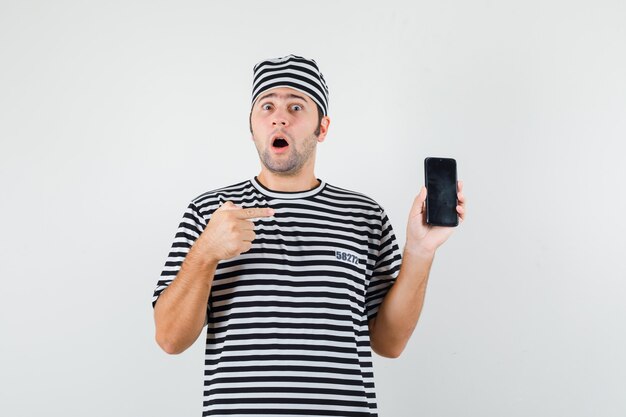 Young male pointing at mobile phone in t-shirt, hat and looking puzzled , front view.
