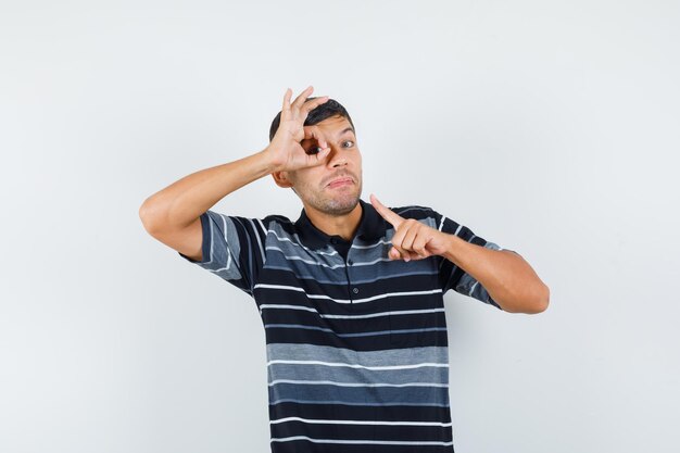 Young male pointing at ok sign on his eye in t-shirt and looking confident. front view.