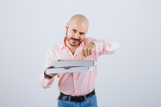 Giovane maschio che indica la scatola della pizza aperta in camicia, jeans e sembra felice. vista frontale.