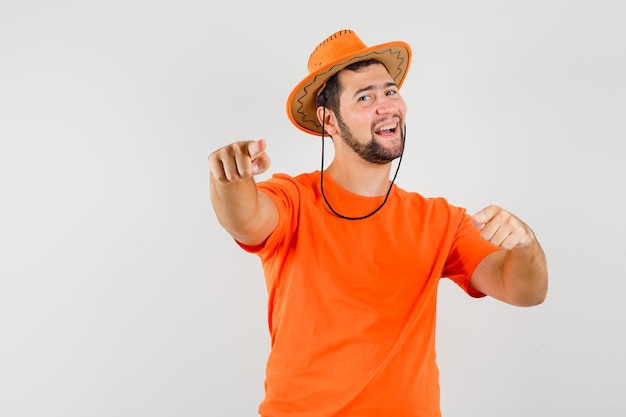 オレンジ色のTシャツ、帽子を指して、陽気に見える若い男性、正面図。