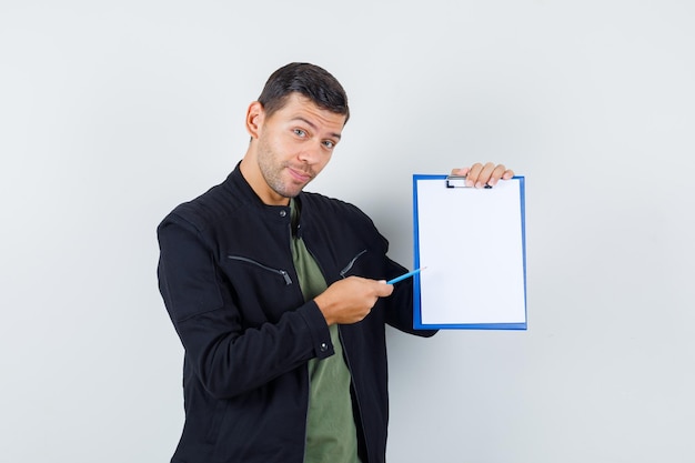 Young male pointing pen at clipboard in t-shirt, jacket and looking cheerful. front view.