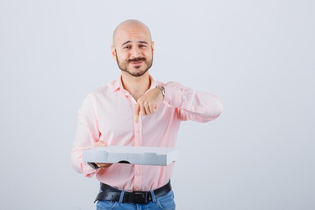 Young male pointing pizza box in shirt, jeans and looking positive. front view.