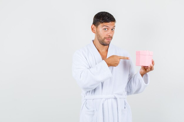 Young male pointing at present box in white bathrobe and looking curious , front view.