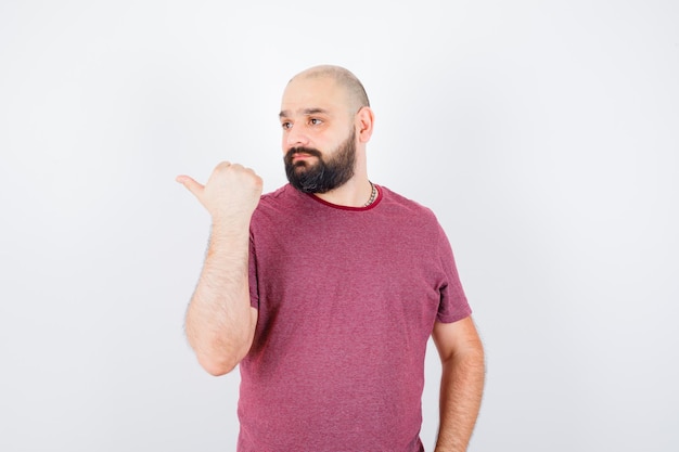 Молодой мужчина, указывая на правую сторону большим пальцем в футболке и нерешительно глядя, вид спереди.
