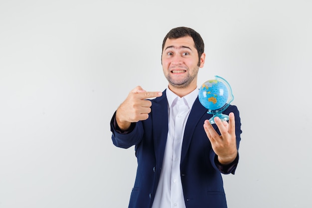 Young male pointing at school globe in shirt and jacket and looking merry