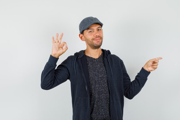 Young male pointing to the side, doing ok sign in t-shirt, jacket, cap and looking cheery , front view.