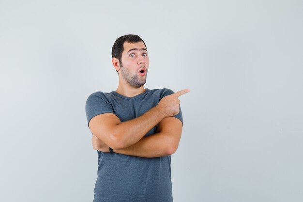 Young male pointing to the side in grey t-shirt and looking surprised. front view.