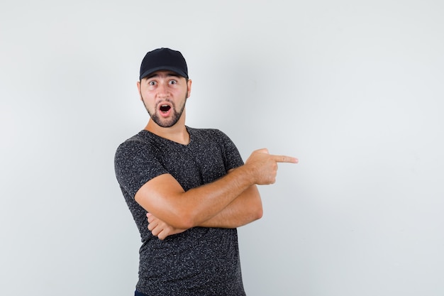 Young male pointing to side in t-shirt and cap and looking curious