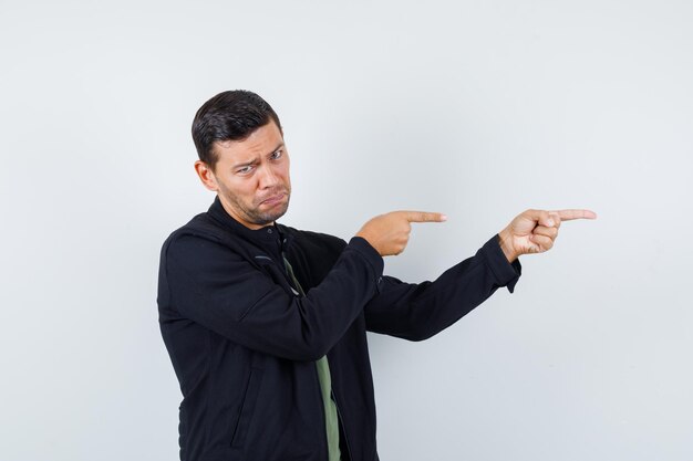 Young male pointing to side in t-shirt, jacket and looking disappointed , front view.