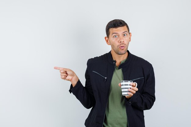 Young male pointing to side while holding cup in t-shirt, jacket and looking puzzled , front view.