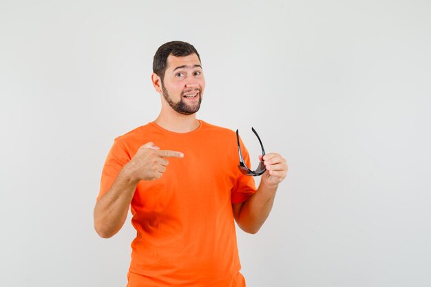 Young male pointing at sunglasses in orange t-shirt and looking confident. front view.