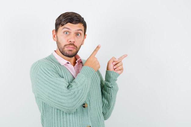 Young male pointing at upper right corner in shirt, cardigan and looking puzzled , front view.