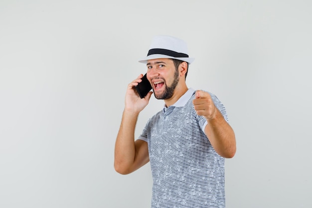 Young male pointing while talking on mobile phone in t-shirt, hat and looking cheery , front view.