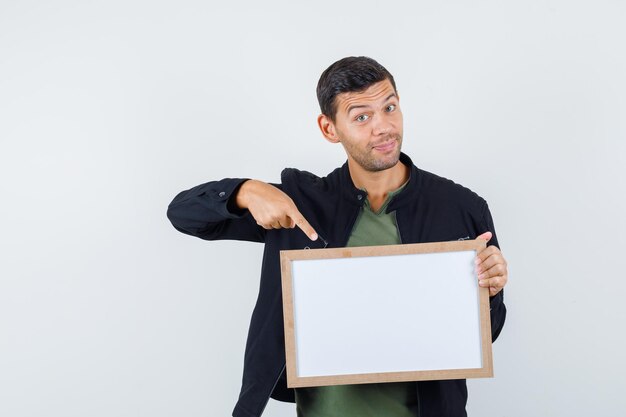 Young male pointing at white board in t-shirt, jacket and looking merry , front view.